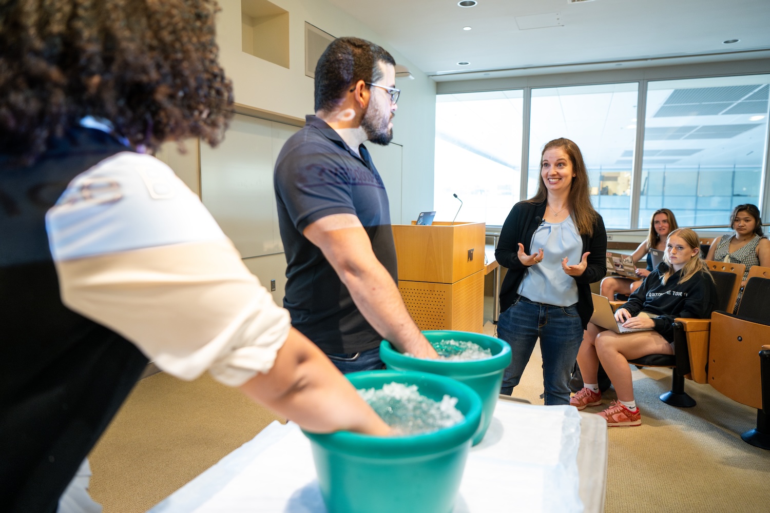 Two students have hands in ice buckets while an instructor speaks to them during a class demonstration