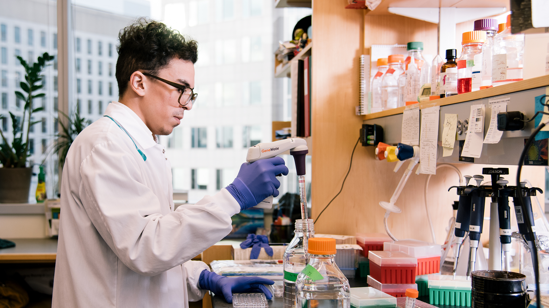 A young man wearing a lab coat and goggles works at a lab bench