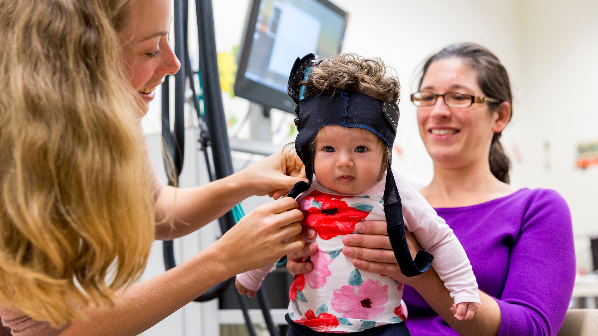 A toddler wearing a headband is held by two smiling female graduate students